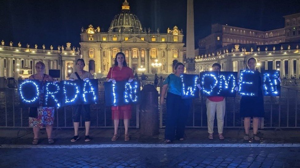 Mujeres de la Conferencia para la Ordenación de Mujeres (WOC) con un cartel de luces que dice "Ordenen mujeres"